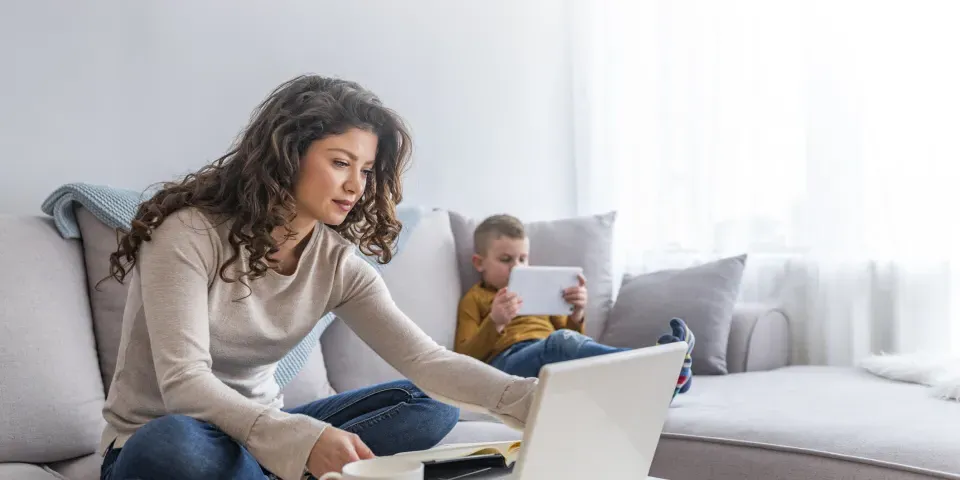 Mother seated on couch taking an online class with son reading on a tablet next to her